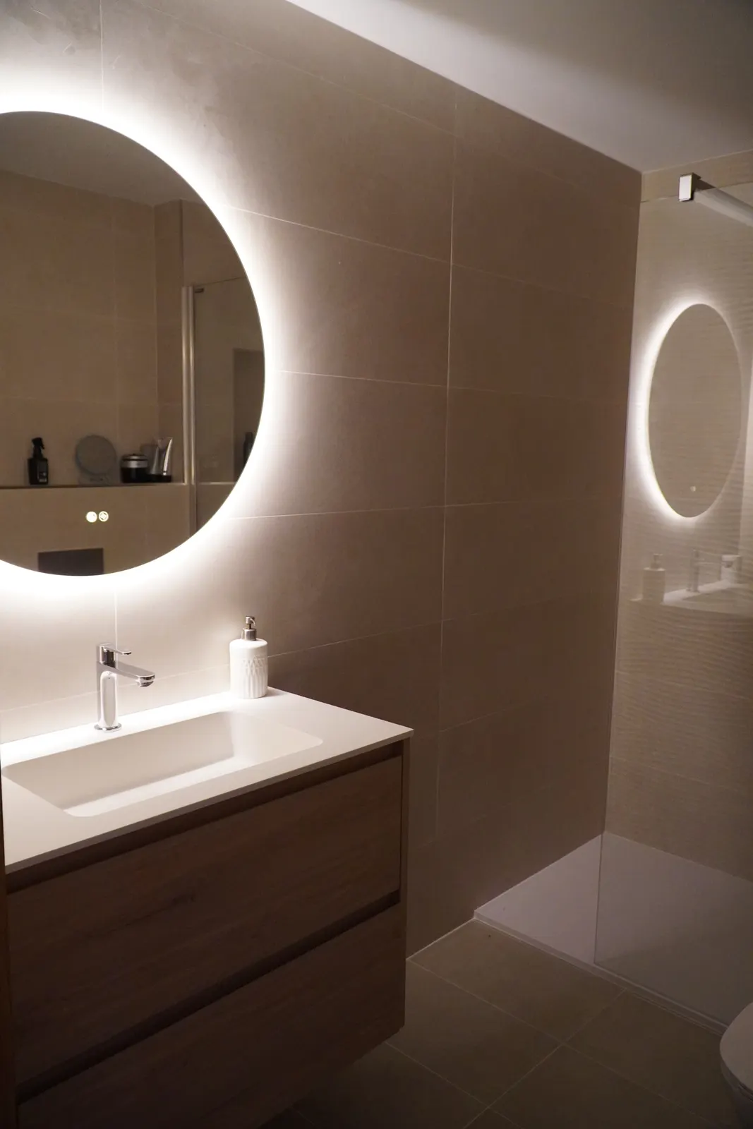 Baño reformado con espejo redondo retroiluminado y paredes de microcemento beige